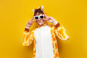 young joyful guy in funny baby giraffe pajamas and glasses is dancing on yellow background