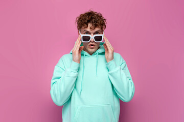 young curly shocked guy in mint hoodie with sunglasses looks in surprise on pink isolated background