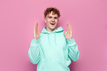 young curly guy in mint hoodie holds empty hands in front of him and shows the size on pink...