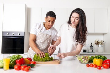 multiracial young couple preparing veggie vegetable and greens salad in white modern kitchen