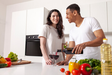 young happy multiracial couple drinking wine in white modern kitchen, african american guy pouring red wine