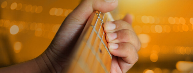 A man's hand is holding a guitar chord against a background of orange lights to convey the...