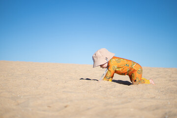 Baby crawls by self on golden sand on hit sunny day with clear sky.
