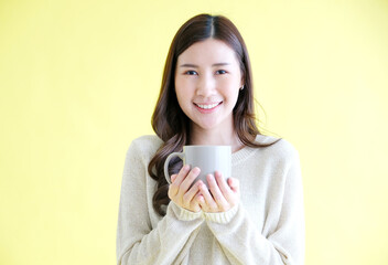 Young asian woman holding coffee cup smiling and looking at camera while standing over isolated yellow background