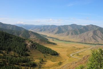 Fototapeta premium A narrow asphalt road windingly crosses a valley sandwiched in the mountains on a sunny summer day.