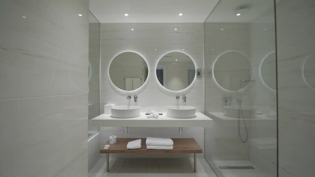 A interior gimbal shot of a beautiful elegant glassy hotel bathroom full of reflections inside of a resort apartment with two round mirrors and white porcelain sinks.