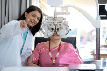 Expert is testing the vision test with diagnostic ophthalmology equipment, Indian or Nepali senior female customer looking through Optical Phoropter during eye exam