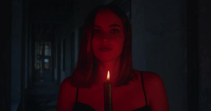 Attractive young woman looking seductively at the camera while holding a candle in a hallway with dim red light.