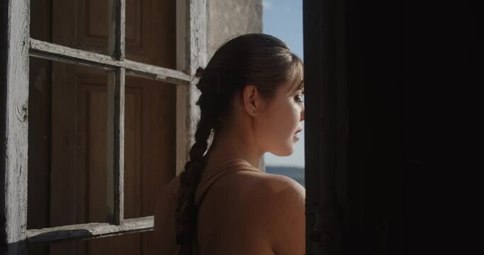 Young brunette woman being caught looking out the window of an old house on a sunny day.