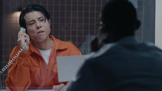 Asian prisoner in orange jumpsuit looking through the glass wall at a photo held by detective, speaking with him on the phone in the visiting room during interrogation