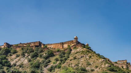 The fortress wall is built on the ridge of the mountain. The watchtower on the background of the blue sky. Green vegetation on the slope. India. Jaipur. Amber Fort