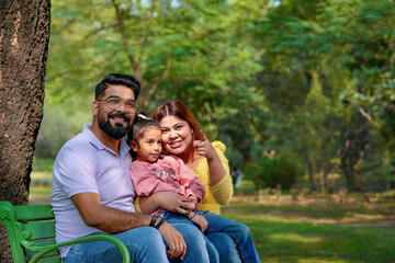 Happy Young indian parents with their cute little daughter sitting at park or garden.
