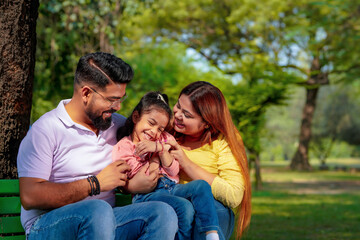 Naklejka premium Happy Young indian parents with their cute little daughter sitting at park or garden.
