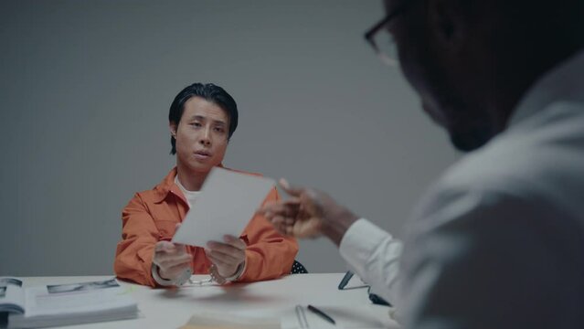 Asian criminal in handcuffs and jail uniform looking at evidence photos, denying his guilt during the interview with black detective in interrogation room. Over the shoulder shot