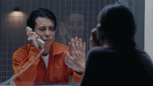 Asian prisoner in orange jumpsuit speaking on the phone and touching hands through glass with loving wife in visiting room