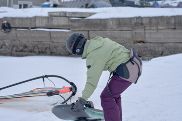A middle-aged woman, a snowsurfer, puts her snowboard on the snow. Preparing to ride a sailboard in the snow on a cloudy winter day.