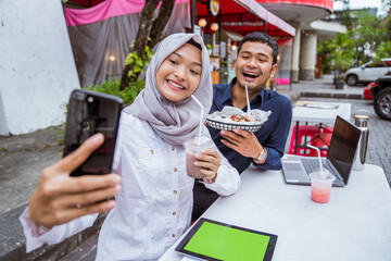 young Muslim woman and boyfriend take a selfie together while breaking the fast drinks and eats at a coffee shop