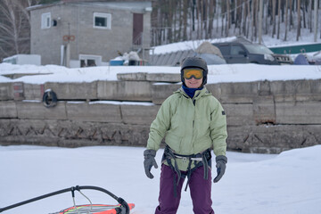 A middle-aged woman, a snowsurfer, stands and waits. Preparing to ride a sailboard in the snow on a cloudy winter day.