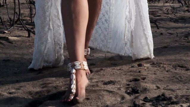 Woman walks barefoot through burnt earth with shell decorations