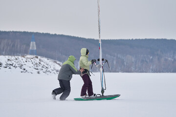 A man, a snowsurfer, helps a woman, his wife, gain speed while moving on a sailboard. A middle-aged man and woman go snowsurfing on a cloudy winter day.