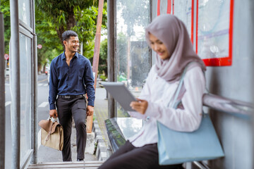 young Asian man walking closer as veiled woman sits with tablet waiting for bus at bus stop