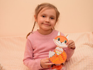 The child learns to embroider with a cross. A cute little girl with pigtails learns to cross-stitch using a safe plastic needle and a cardboard trainer in the shape of a fox or a kitten.