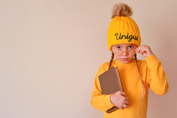 A unique child. A little cute girl in an orange hat with the inscription Unique and glasses, holds a book and looks attentively. Smart, educated, intelligent child. Love of books. Education children