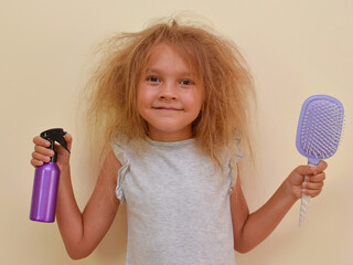 A cute little girl with very tangled hair holds a spray can with hair conditioner and a massage...