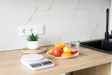 A plate of fresh fruit on the kitchen table, a kitchen scale and a glass of water near the sink. The concept of healthy and proper nutrition