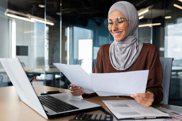 Successful muslim woman in hijab working inside office with documents, joyful financial accountant happy with reports checking financial data and smiling, businesswoman at workplace with laptop.