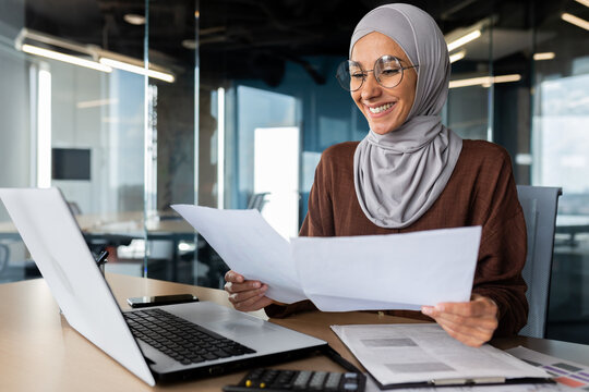 Successful muslim woman in hijab working inside office with documents, joyful financial accountant happy with reports checking financial data and smiling, businesswoman at workplace with laptop.