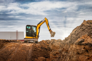 Mini excavator at the construction site on the edge of a pit against a cloudy blue sky. Compact...