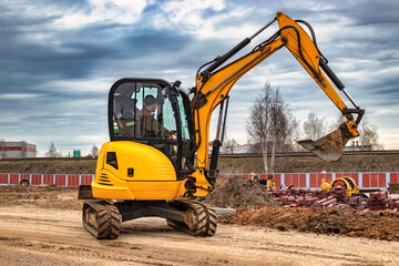 Mini excavator at the construction site on the edge of a pit against a cloudy blue sky. Compact...