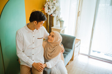 romantic married Asian Muslim couple looking at each other while sitting on sofa together in the living room