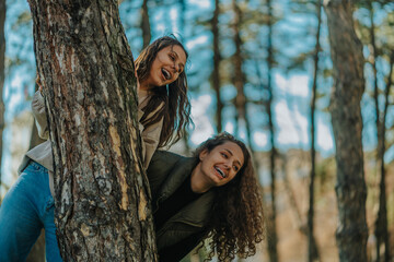 Young stunning girls playing and having fun outdoors. They are looking away and smiling while hiding behind tree in the park