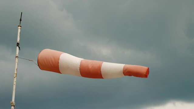 A white and red windsock on a pipe with peeling paint ripples in the wind. In the background is a dark sky with a cloud. Close-up. Slow motion