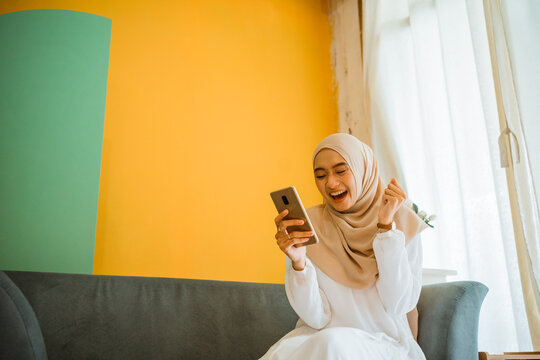 portrait of excited Asian veiled woman sitting on couch using cell phone at home