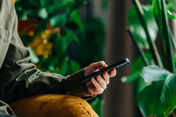Woman in green shirt holds smartphone in hand on palm leaves background and looking in to display. Side view with plants in bokeh