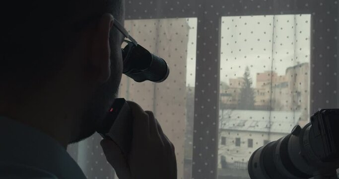 Detective uses binoculars and camera to observe suspect on street through window curtain in darkened room