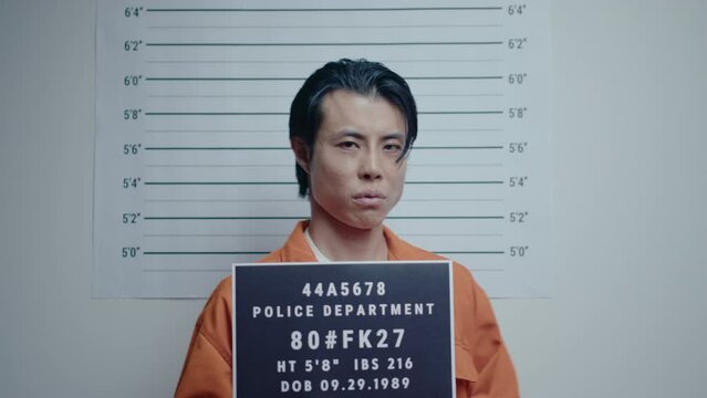Mugshot of Asian criminal in orange uniform holding a sign posing on camera against height chart, taking photos of suspect after arrest in police department
