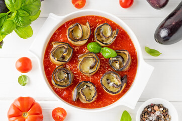 Eggplant rolls with cheese in tomato sauce in a white ceramic baking form on a white wooden background. Vegetarian healthy dish. Selective focus, top view.