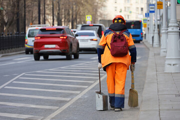 Woman worker in orange uniform with a broom sweeps the road on cars background. Street cleaning in spring city