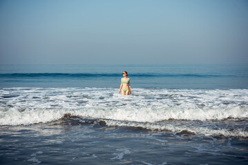 A girl in a swimsuit stands in the waves on the seashore and enjoys swimming and relaxing in a warm country. General view, space for text