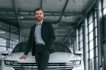 Man in black suit is indoors in the car dealership near automobile