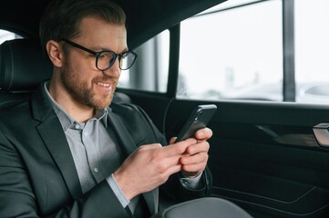 Holding smartphone. Man in formal business clothes is sitting in the modern automobile