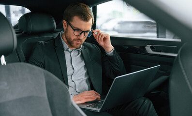 Fototapeta na wymiar With laptop, working. Man in formal business clothes is sitting in the modern automobile