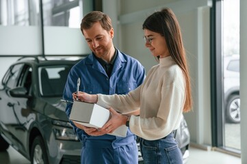 Obraz premium Man is holding box and notepad. Young woman is visiting auto mechanic in garage. Repair service