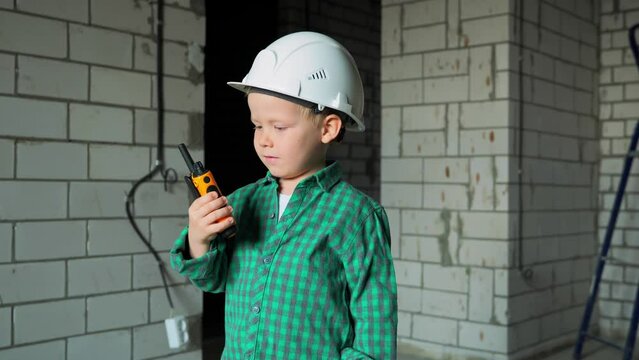 A 5-year-old boy of the buzzer Z generation plays on a construction site in a white helmet and with a walkie-talkie in his hand. A boy in a green shirt is talking on a radio at a site. High quality 4k