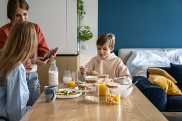 Obraz na płótnie Canvas Mother and daughter using mobile phone at kitchen table while having breakfast together with family. Teen girl showing mom something on smartphone during mealtime. Cell phone usage and family bond