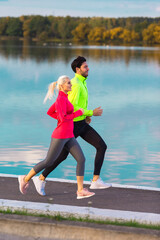 Jogging Ideas. Running Couple While Training Outdoors on Road At River For Durable Marathon Pretty Young Female and Extremely Fit Caucasian Handsome Man.